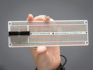 A white hand holding up a Adafruit Perma-Proto Raspberry Pi Breadboard PCB.
