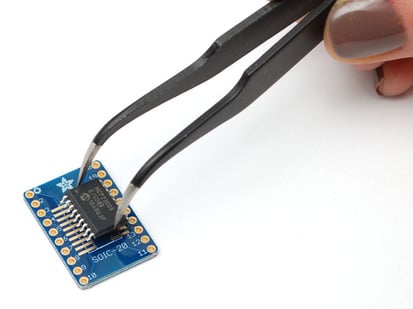 SMT Breakout PCB for SOIC-20 or TSSOP-20 - 3 Pack! - A pair of tweezers holding a SOIC chip hovers over the SMT Breakout PCB for SOIC-20 or TSSOP-20.