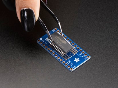 SMT Breakout PCB for SOIC-28 or TSSOP-28 - 3 Pack! - A pair of tweezers holding a microchip hovers over the SMT Breakout PCB for SOIC-28 or TSSOP-28.