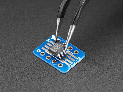 SMT Breakout PCB for SOIC-8, MSOP-8 or TSSOP-8 - 6 Pack! - A pair of tweezers holding a microchip hovers over the SMT Breakout PCB for SOIC-8, MSOP-8 or TSSOP-8.