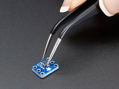SMT Breakout PCB Set For SOT-23, SOT-89, SOT-223 and TO252 - A pair of tweezers holding a microchip hovers over a small rectangular Breakout PCB.