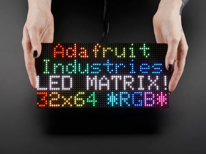 64x32 RGB LED Matrix - 4mm pitch - Two white hands hold out an assembled and powered on 64x32 RGB LED Matrix Panel - 4mm pitch. The matrix displays "Adafruit Industries LED MATRIX! 32x64 *RGB*"
