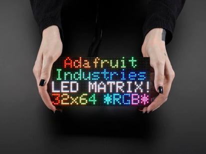 64x32 RGB LED Matrix - 3mm pitch - Two white hands hold out an assembled and powered on 64x32 RGB LED Matrix Panel - 3mm pitch. The matrix displays "Adafruit Industries LED MATRIX! 32x64 *RGB*"