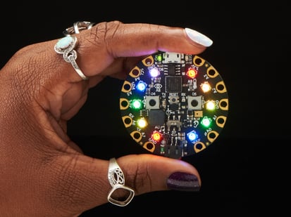 Circuit Playground Express - A Black woman's manicured hand holds a round microcontroller with lit up LEDs.