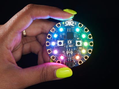 Circuit Playground Bluefruit - Bluetooth® Low Energy - shot of a Black woman's neon-green manicured hand holding up a Circuit Playground Bluefruit glowing rainbow LEDs.
