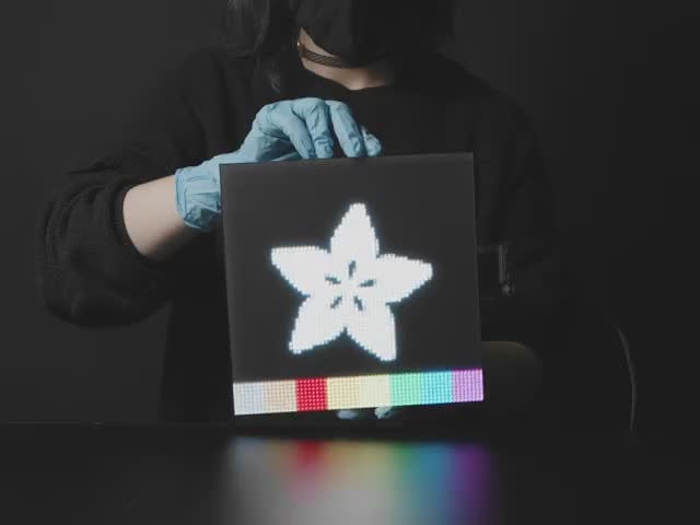 64x64 RGB LED Matrix - 3mm Pitch - 192mm x 192mm - 192mm x 192mm - Video of a woman in black slowly rotating a powered-on 64x64 RGB LED Matrix. The matrix displays the Adafruit star flower logo with rainbow sand falling animation.