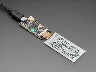 Angled shot of a 2.13" monochromatic electronic ink display module connected to a driver board. The display features a graphic python with the text: 2.13" ThinkInk Mono SSD1680 feather"