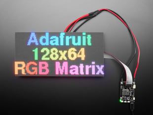 LED matrix panel wired up and displaying "Adafruit 128x64 RGB matrix" in rainbow colors.
