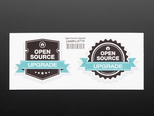 photograph of two black stickers, one shield shaped, the other gear shaped, with white lettering reading "open source" with a turquoise banner running across each reading "upgrade" both stickers are mounted on white paper with barcode. 